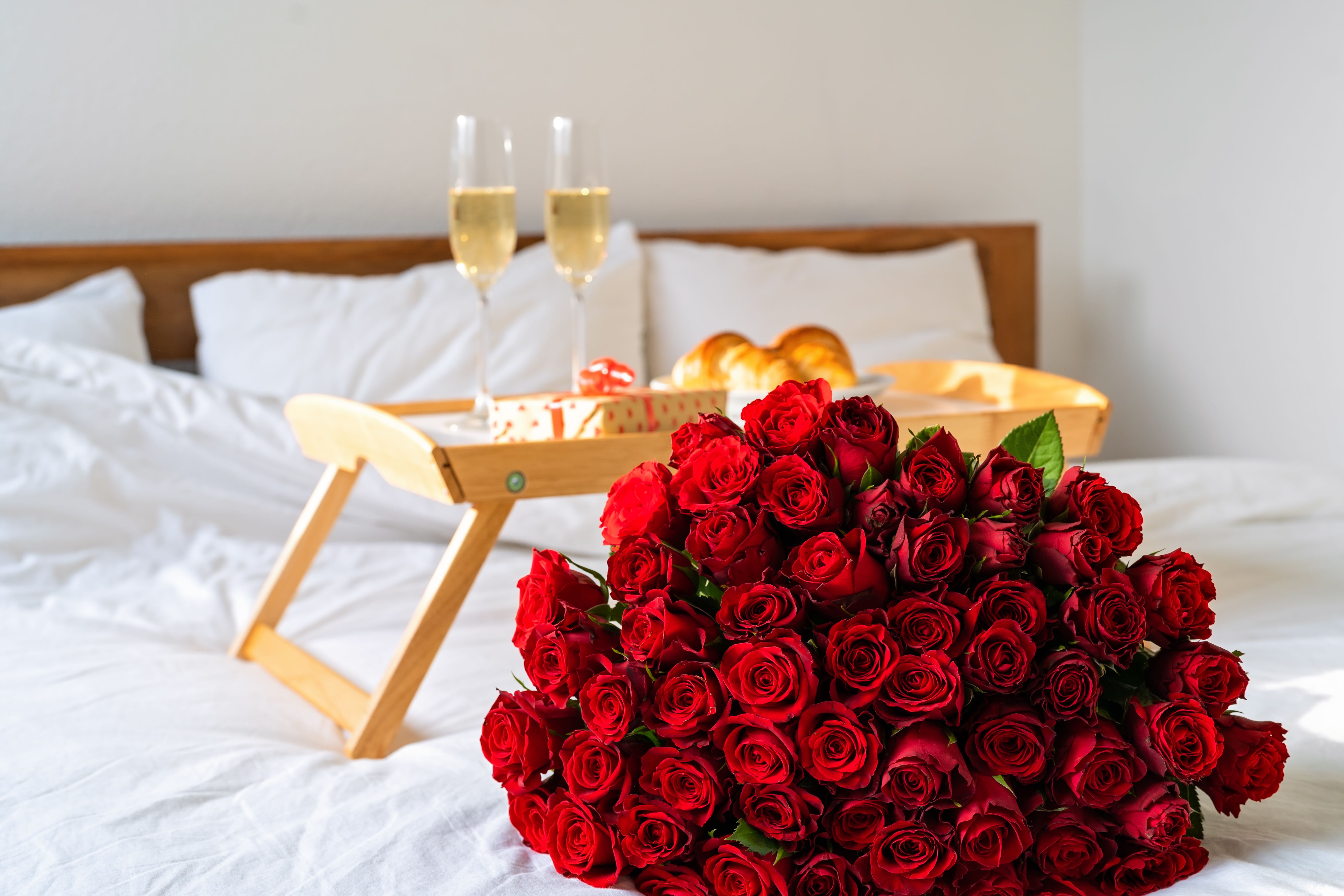 Grand bouquet de roses rouges posé sur un lit, à côté d’un petit-déjeuner pour deux.