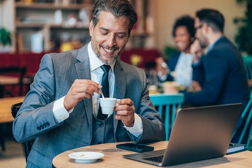 Homme d’une quarantaine d’années souriant dans un café avec un ordinateur portable et une tasse de café, illustrant une rencontre sérieuse, une première impression positive et une communication authentique.