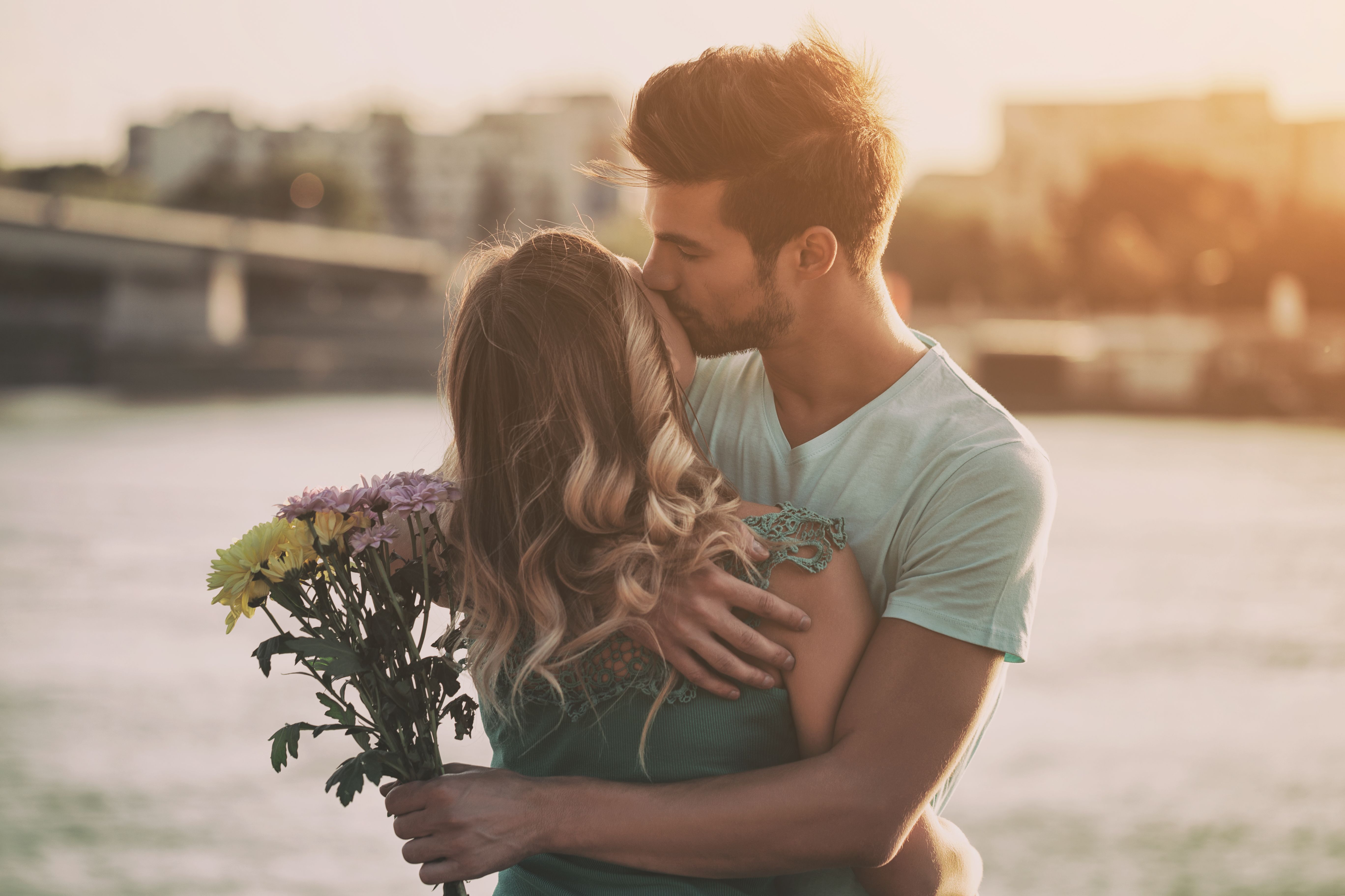 Jeune couple souriant s’enlaçant en ville, l’homme tenant un bouquet de fleurs, illustrant une rencontre sérieuse, une relation durable et un premier rendez-vous réussi.